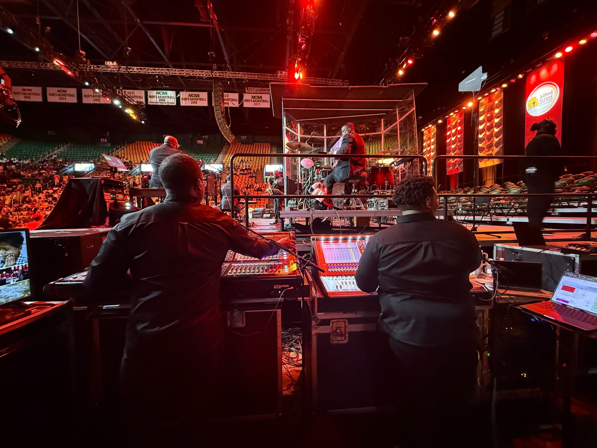 Two engineers working at stage-side mixing consoles, with the band, drum enclosure, and arena beyond them.